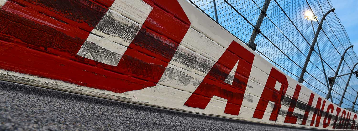 Close-up view of the red and white painted wall at Darlington Raceway, marked with tire scuffs, with safety fencing and track lights overhead under a blue sky.