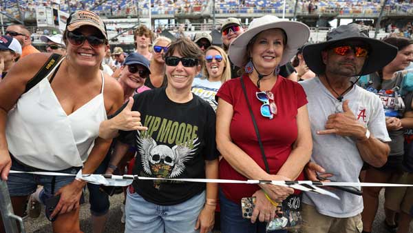 Smiling fans crowd together near a barrier at a NASCAR race event, with grandstands visible in the background on a sunny day.