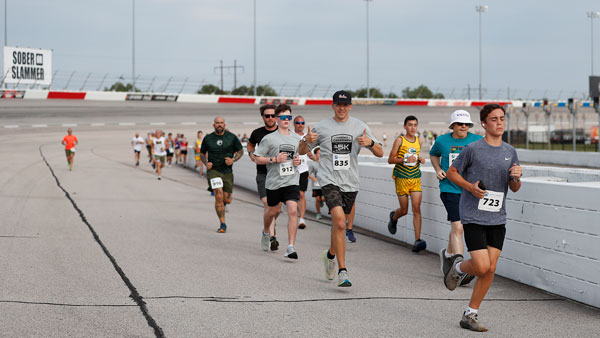 A group of runners of varying ages jog along a wide racetrack during a daytime 5K event. The participants wear athletic clothing such as T-shirts, shorts, and running shoes, with numbered race bibs pinned to their shirts. They are spread out in a line, moving forward along the track surface beside a white retaining wall. In the background, more runners follow, and track signage and fencing are visible. The setting appears to be an active race event, with a casual, community-focused atmosphere.